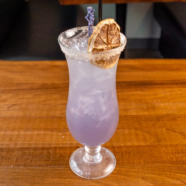 Lavender cocktail with a sugared rim, dried orange slice, and lavender sprig on a wooden table.
