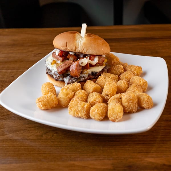 A loaded burger with melted cheese, sausage, and onions, served with a side of tater tots.