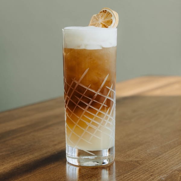 A tall, etched glass with a layered brown and white drink, topped with foam and a dried orange slice.