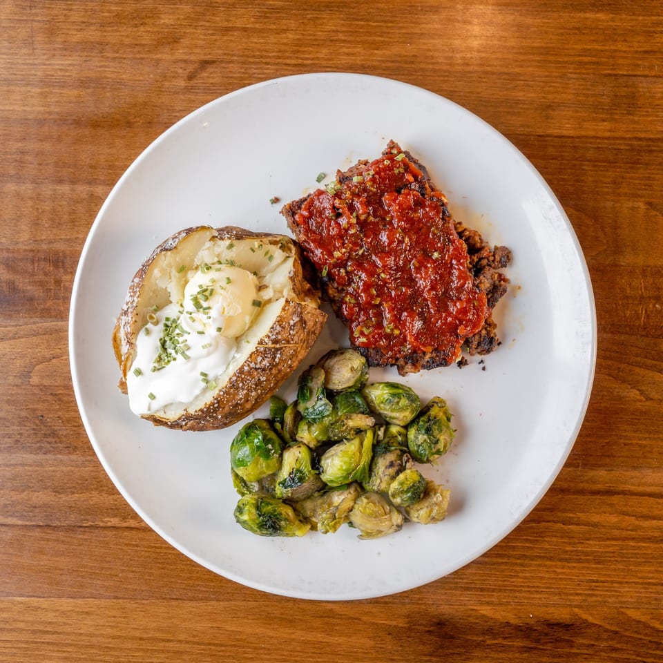 Meatloaf with tomato sauce, a baked potato with sour cream and chives, and roasted Brussels sprouts.