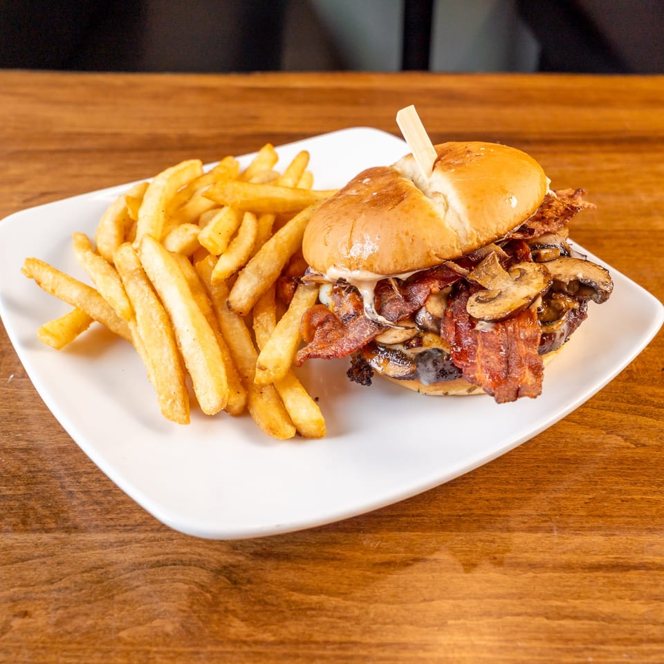 A gourmet burger with bacon and mushrooms, served with a side of golden french fries on a white plate.