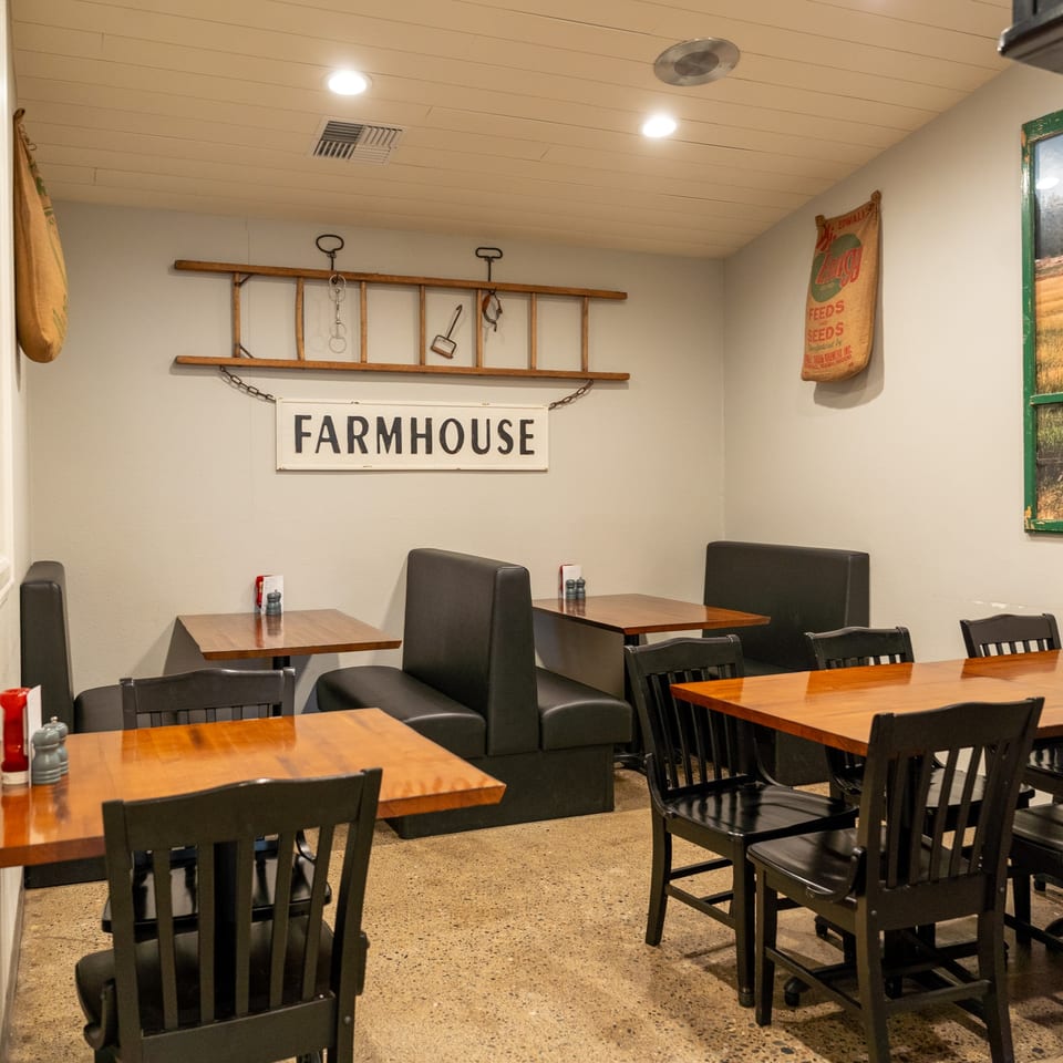Restaurant dining room with wooden tables, black chairs, and booths, featuring a "FARMHOUSE" sign.