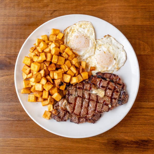A white plate holds a steak with melting butter, crispy home fries, and two fried eggs.