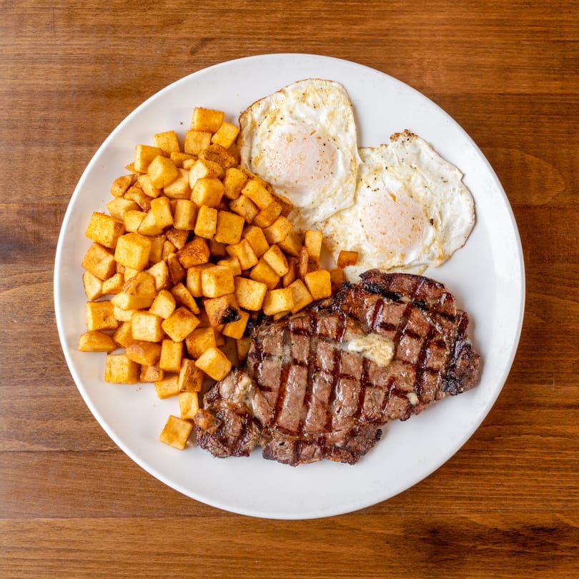 A white plate holds a steak with melting butter, crispy home fries, and two fried eggs.
